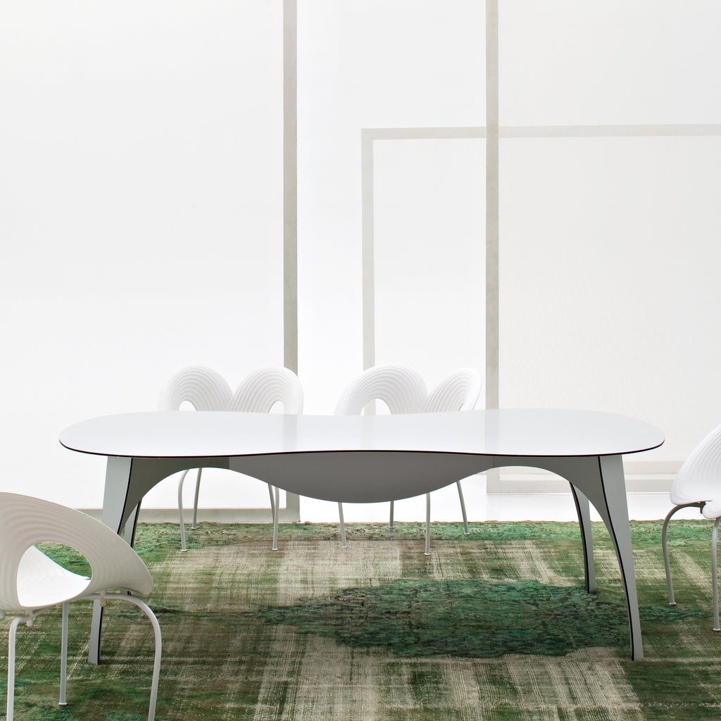 An oval white dining table with curved pedestal legs, surrounded by white chairs in a bright showroom.